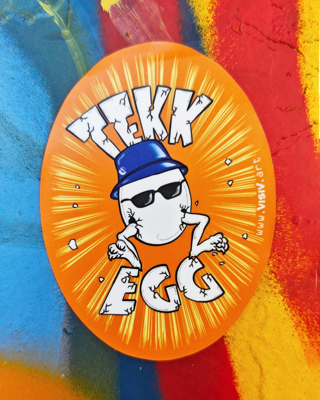 Orange oval Tekk Egg sticker photographed on a painted wall with blue, red and yellow brushstroke textures, featuring a cartoon egg wearing a blue cap and sunglasses.